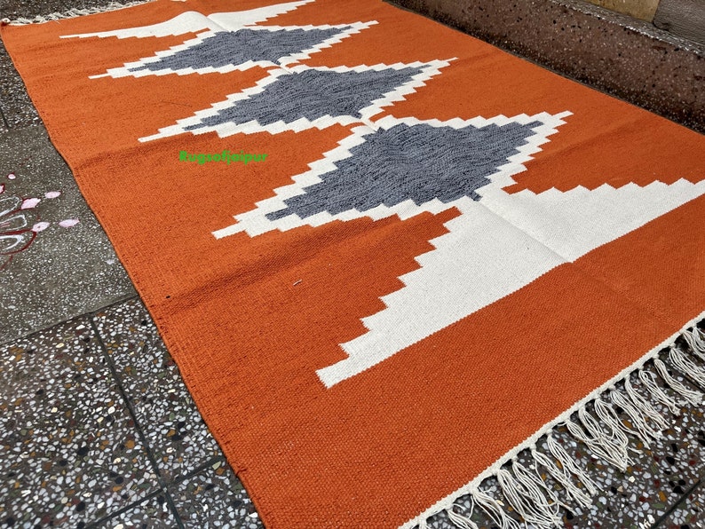 Puede incluir: Alfombra geom&eacute;trica con estampado naranja, blanco y gris con flecos blancos.