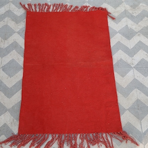 May include: A rectangular red rug with fringed edges. The rug is made of a woven material and is placed on a gray and white patterned tile floor. The rug's color is a vibrant, solid red.