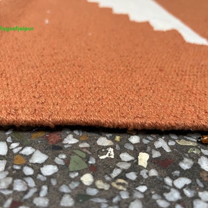 Puede incluir: Primer plano del borde de una alfombra naranja quemada con un patr&oacute;n geom&eacute;trico blanco. La alfombra est&aacute; sobre una superficie texturizada con peque&ntilde;as piedras de colores.