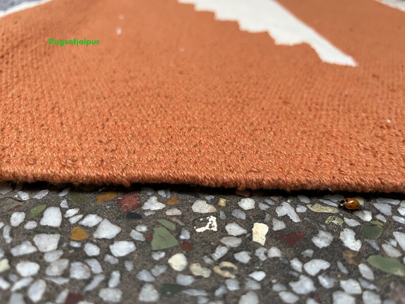 Puede incluir: Primer plano del borde de una alfombra naranja quemada con un patr&oacute;n geom&eacute;trico blanco. La alfombra est&aacute; sobre una superficie texturizada con peque&ntilde;as piedras de colores.