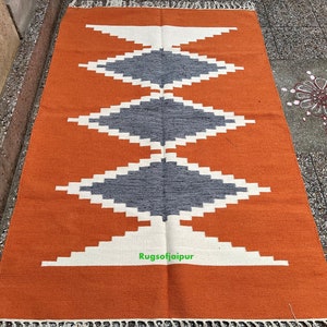 Puede incluir: Una alfombra tejida a mano con un patr&oacute;n geom&eacute;trico en tonos naranja, blanco y gris. La alfombra tiene un borde de flecos y est&aacute; hecha de fibras naturales.