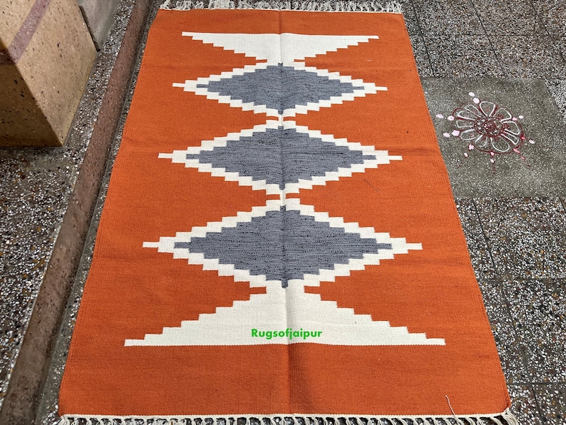 Puede incluir: Una alfombra tejida a mano con un patr&oacute;n geom&eacute;trico en tonos naranja, blanco y gris. La alfombra tiene un borde de flecos y est&aacute; hecha de fibras naturales.