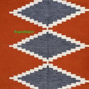 Puede incluir: Primer plano de una alfombra tejida con un patr&oacute;n geom&eacute;trico. La alfombra tiene una forma de diamante gris oscuro sobre un fondo blanco, con un fondo de color &oacute;xido. El texto "Rugsofjaipur" es visible en la imagen.