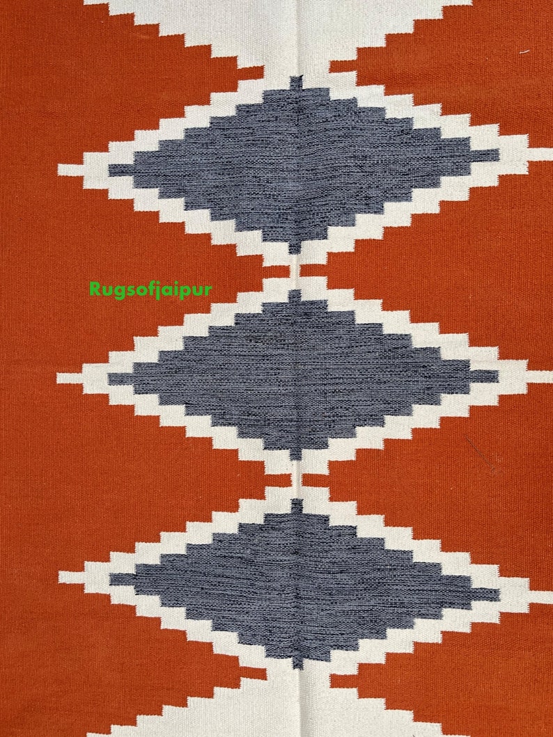 Puede incluir: Primer plano de una alfombra tejida con un patr&oacute;n geom&eacute;trico. La alfombra tiene una forma de diamante gris oscuro sobre un fondo blanco, con un fondo de color &oacute;xido. El texto "Rugsofjaipur" es visible en la imagen.