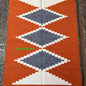 Puede incluir: Alfombra geom&eacute;trica de color naranja, blanco y gris con flecos. La alfombra tiene un patr&oacute;n de diamantes con un borde blanco y un centro gris. El texto "Rugsofjaipur" es visible en la esquina inferior izquierda.
