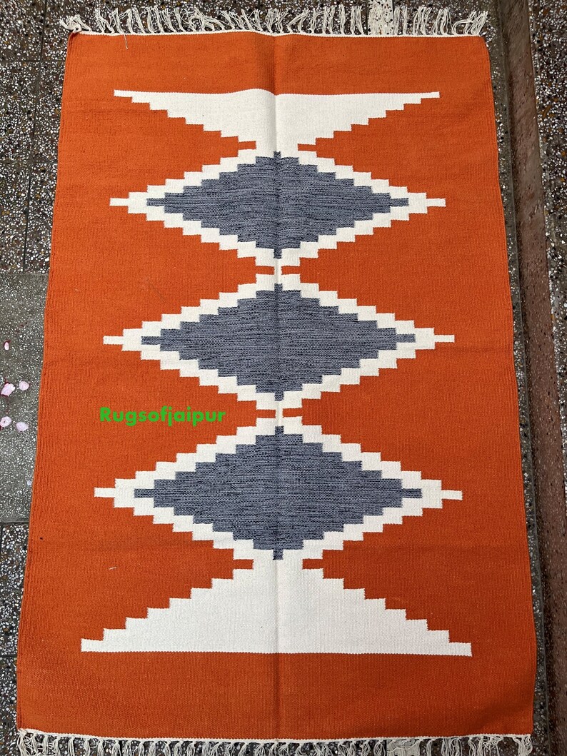 Puede incluir: Alfombra geom&eacute;trica de color naranja, blanco y gris con flecos. La alfombra tiene un patr&oacute;n de diamantes con un borde blanco y un centro gris. El texto "Rugsofjaipur" es visible en la esquina inferior izquierda.