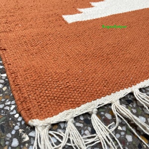 Puede incluir: Primer plano de una alfombra tejida de color naranja terracota con flecos blancos. La alfombra tiene un patr&oacute;n geom&eacute;trico con rayas blancas.