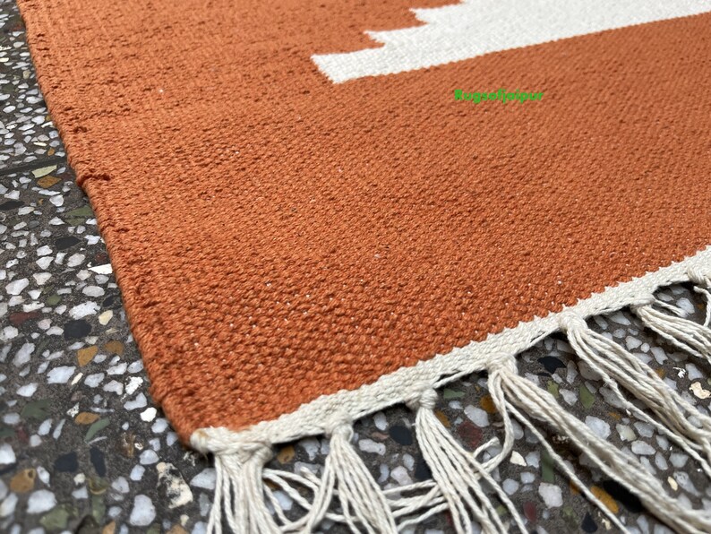 Puede incluir: Primer plano de una alfombra tejida de color naranja terracota con flecos blancos. La alfombra tiene un patr&oacute;n geom&eacute;trico con rayas blancas.