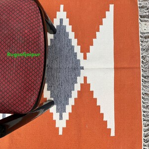 Puede incluir: Una alfombra tejida a mano con un patr&oacute;n geom&eacute;trico en tonos de blanco, gris y naranja. La alfombra tiene un borde de flecos y est&aacute; hecha de algod&oacute;n. El texto "Rugsofjaipur" es visible en la esquina inferior izquierda de la imagen.