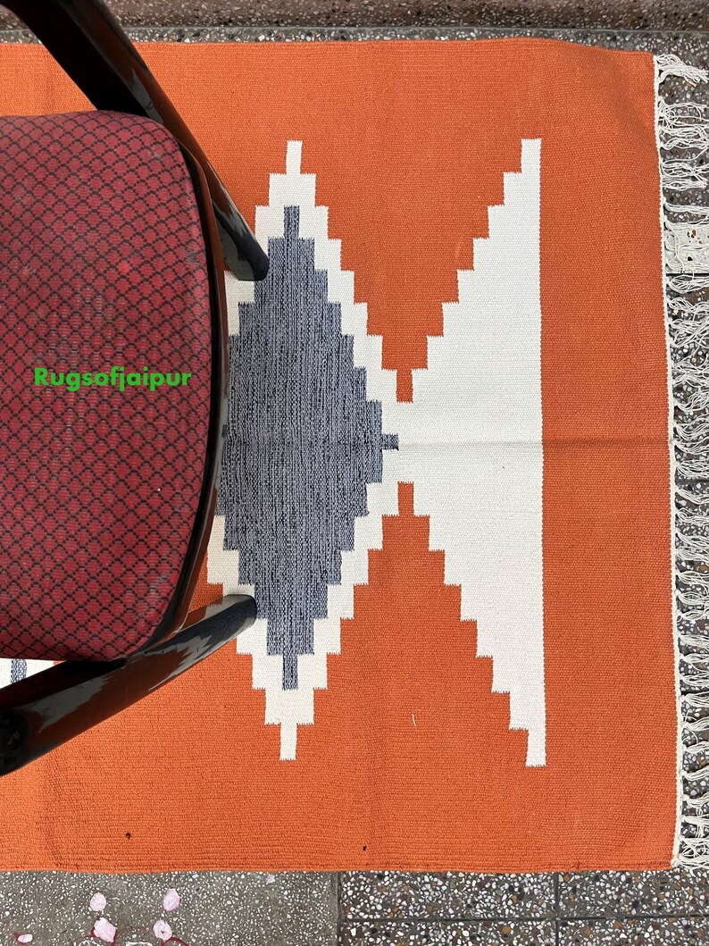 Puede incluir: Una alfombra tejida a mano con un patr&oacute;n geom&eacute;trico en tonos de blanco, gris y naranja. La alfombra tiene un borde de flecos y est&aacute; hecha de algod&oacute;n. El texto "Rugsofjaipur" es visible en la esquina inferior izquierda de la imagen.