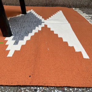 Puede incluir: Primer plano de una alfombra de color &oacute;xido con un patr&oacute;n geom&eacute;trico en blanco y gris. La alfombra tiene un borde de flecos.