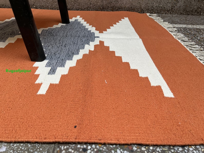 Puede incluir: Primer plano de una alfombra de color &oacute;xido con un patr&oacute;n geom&eacute;trico en blanco y gris. La alfombra tiene un borde de flecos.