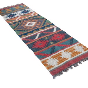 May include: A long, narrow rug with a colorful geometric pattern in shades of red, orange, blue, green, and white. The rug has a fringe on both ends.