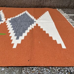 Puede incluir: Primer plano de una alfombra tejida con un patr&oacute;n geom&eacute;trico en tonos naranja, blanco y gris. La alfombra tiene un borde de flecos.