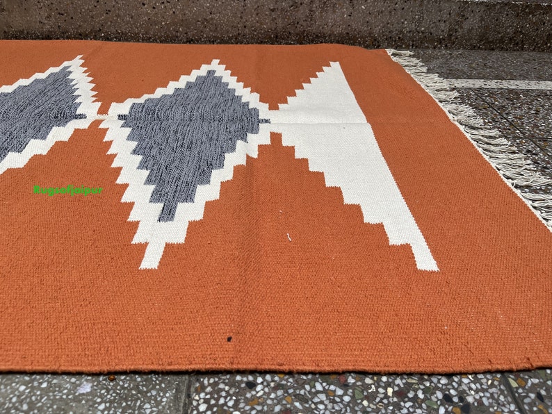 Puede incluir: Primer plano de una alfombra tejida con un patr&oacute;n geom&eacute;trico en tonos naranja, blanco y gris. La alfombra tiene un borde de flecos.