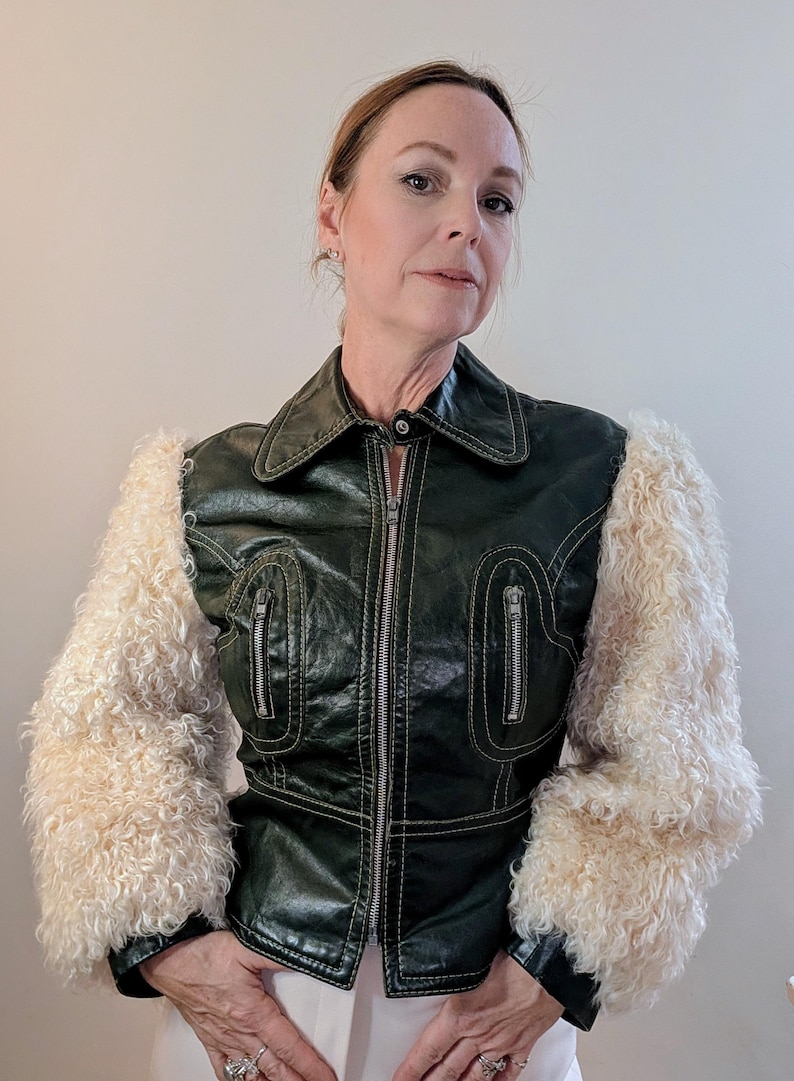 May include: A dark green jacket with cream-colored, fluffy sleeves. The jacket has a zipper closure, two zippered pockets, and white stitching. The collar is a classic style.