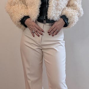 May include: Cream-colored faux fur jacket with a black leather-look collar and cuffs. The jacket is paired with cream-colored, wide-leg trousers. The outfit is a stylish ensemble.
