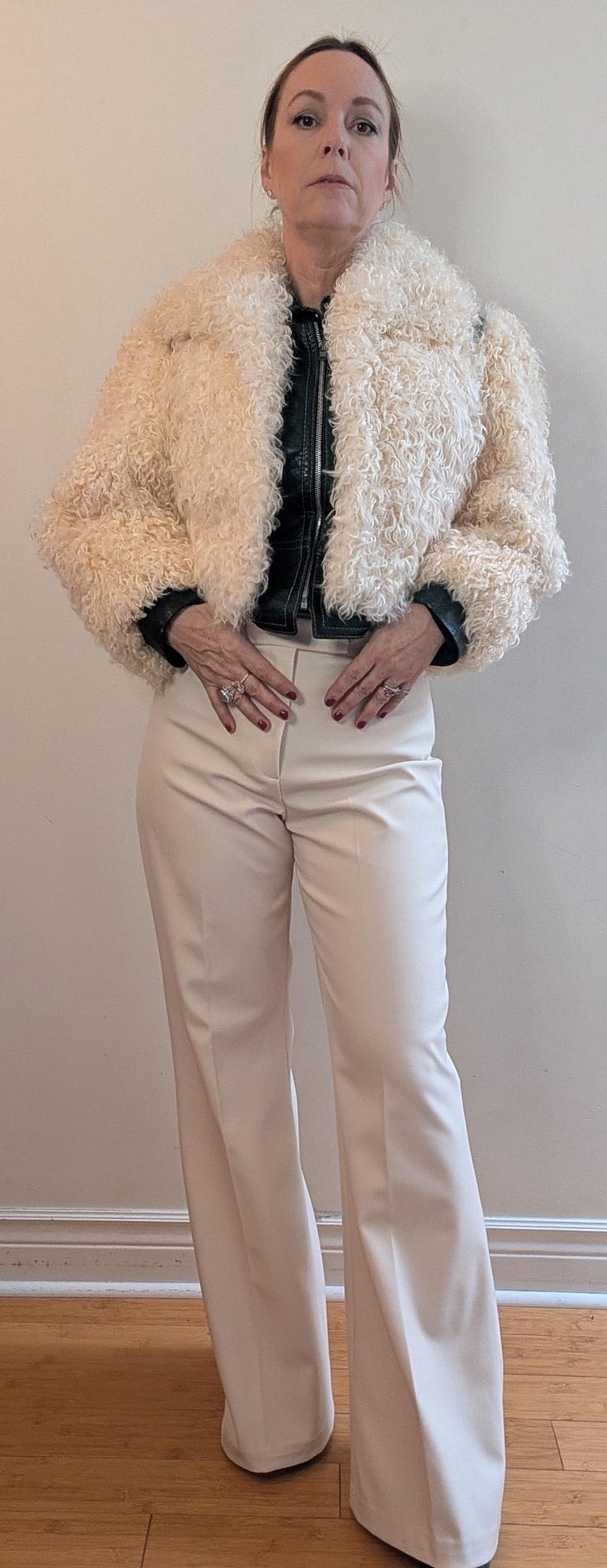 May include: Cream-colored faux fur jacket with a black leather-look collar and cuffs. The jacket is paired with cream-colored, wide-leg trousers. The outfit is a stylish ensemble.