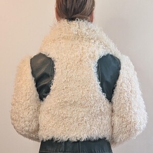 May include: A cream-colored, curly shearling jacket with black leather accents on the shoulders and lower back. The jacket has a high collar and a cropped length, offering a unique texture and style.