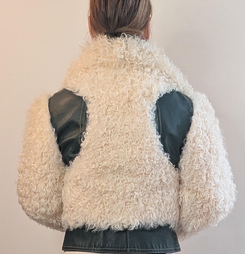 May include: A cream-colored, curly shearling jacket with black leather accents on the shoulders and lower back. The jacket has a high collar and a cropped length, offering a unique texture and style.