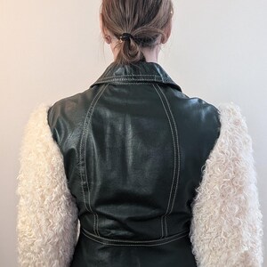 May include: A dark green faux leather jacket with cream-colored, fluffy sleeves. The jacket has white stitching details and a classic collar. The model is wearing white pants. The jacket is a unique fashion statement.