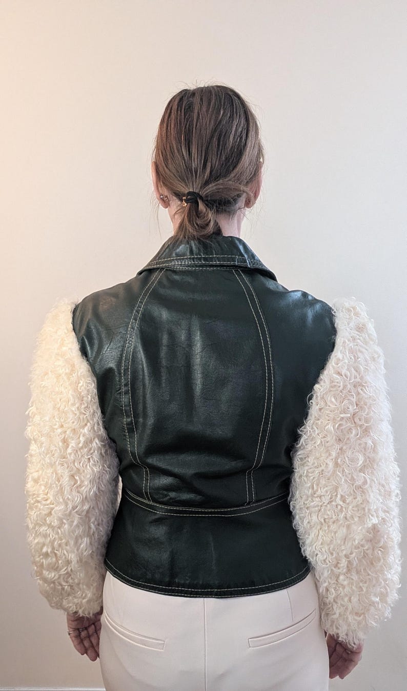 May include: A dark green faux leather jacket with cream-colored, fluffy sleeves. The jacket has white stitching details and a classic collar. The model is wearing white pants. The jacket is a unique fashion statement.