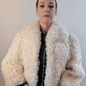 May include: Cream-colored, curly faux fur jacket with a wide collar and a black leather-like zipper. The jacket has a cropped design and is worn over a black top. The model is wearing light-colored pants.