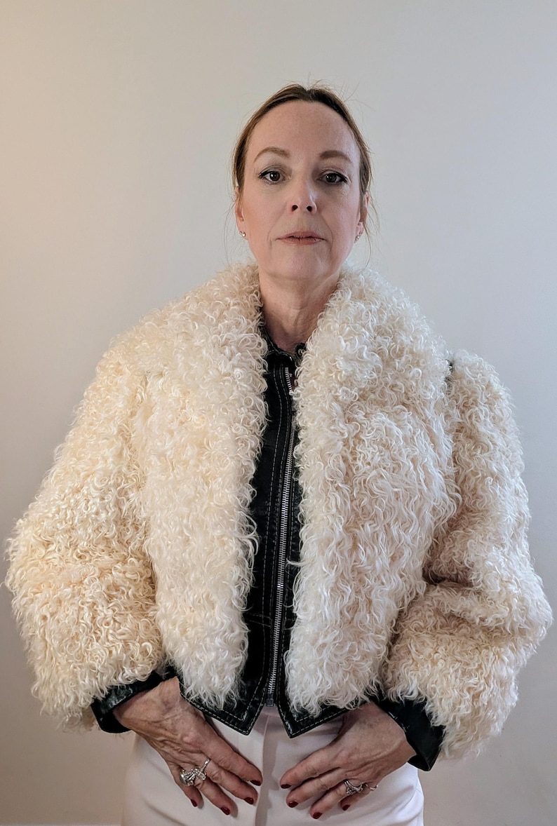 May include: Cream-colored, curly faux fur jacket with a wide collar and a black leather-like zipper. The jacket has a cropped design and is worn over a black top. The model is wearing light-colored pants.