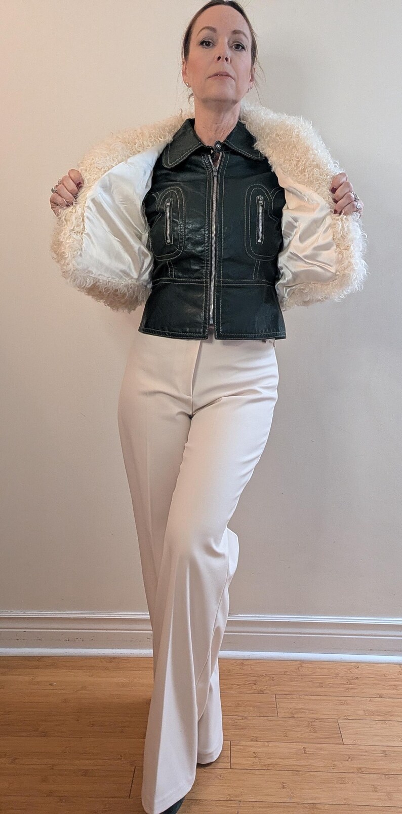 May include: A vintage-style outfit featuring a dark green leather jacket with a cream-colored shearling collar and lining, paired with cream-colored flared pants. The jacket has a zipper closure and decorative stitching.