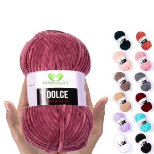 Op de afbeelding: Een bol zacht, roze fluweelgaren genaamd "Dolce" van Meriwoolart. Het garen bevindt zich in een hand, en er zijn 12 andere kleur opties te zien op de achtergrond.