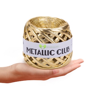 Peut inclure: Une pelote de fil métallique doré, portant l'inscription "METALLIC CLUB" en lettres noires. Le fil est tissé de manière serrée, et l'étiquette comprend également le logo "MERIWOOLART". La pelote est tenue dans une main sur fond blanc.