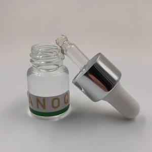 May include: A clear glass bottle with a silver and white dropper top. The bottle has a green and gold label with the word "ANOQ" on it.