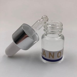 Puede incluir: Una botella de vidrio transparente con un tapón cuentagotas plateado. La botella tiene una etiqueta azul y dorada con la palabra "ANO" en ella.