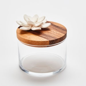 Op de afbeelding: Een glazen pot met een houten deksel met daarop een witte keramische bloem. Het deksel is gemaakt van twee verschillende soorten hout.