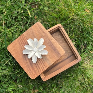 May include: A wooden box with a hinged lid. The lid has a white ceramic flower design on top. The box is made of dark wood with a natural grain.