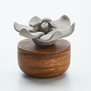 May include: A gray ceramic flower with five petals sits atop a round wooden base. The flower has a textured surface and a small, raised center.