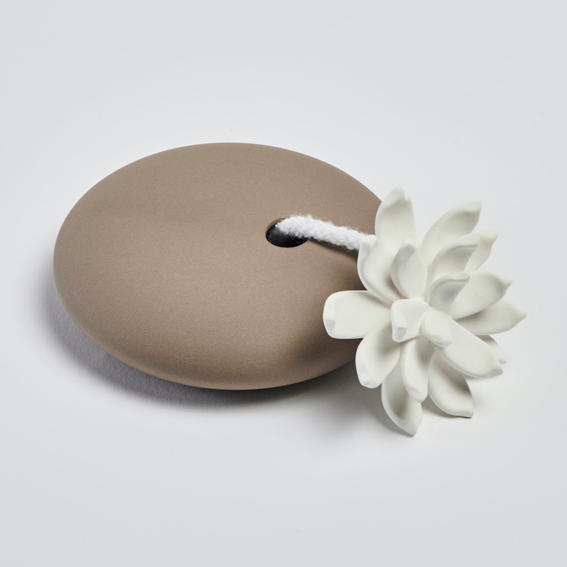 May include: A brown ceramic diffuser with a white flower-shaped diffuser stick. The stick is attached to the diffuser with a white string.