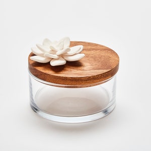 Op de afbeelding: Een heldere glazen pot met een houten deksel. Een witte keramische vetplantbloem versiert de bovenkant van het deksel. De pot is rond en staat op een wit oppervlak. Het houten deksel heeft een warme bruine kleur.