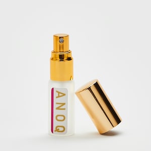 Könnte beinhalten: Eine kleine, klare Glasflasche mit goldener Kappe und goldener Sprühdüse. Die Flasche hat ein weißes Etikett mit dem Wort "ANOQ" in goldenen Buchstaben. Die Flasche steht auf einer weißen Oberfläche.