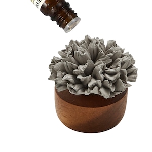 May include: A gray ceramic flower diffuser with a wooden base. An essential oil bottle is being poured onto the diffuser.