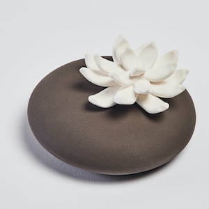 Può includere: Una pietra diffusore in ceramica grigio scuro con un fiore in ceramica bianca in cima. Il fiore ha più petali ed è a forma di loto.