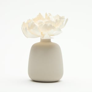 Könnte beinhalten: Eine kleine, cremefarbene Keramikvase mit rundem Körper und schmalem Hals, die eine große, cremefarbene Papierblume enthält. Die Blume hat mehrere Schichten zarter Blütenblätter. Die Vase ist etwa 10 cm hoch.