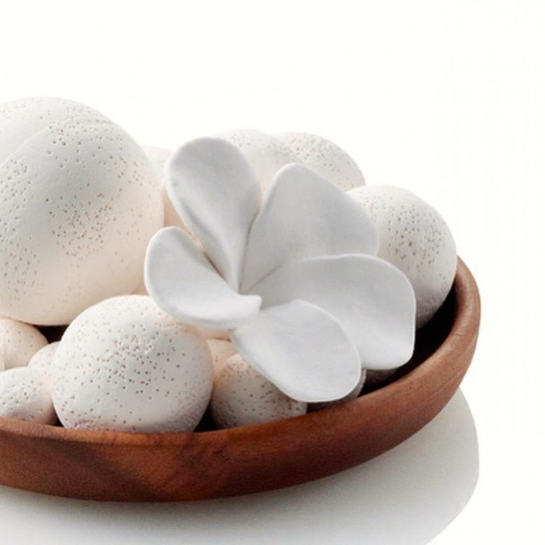 May include: A white ceramic flower with five petals sits in a wooden bowl filled with white ceramic balls. The flower is a decorative element for a home.