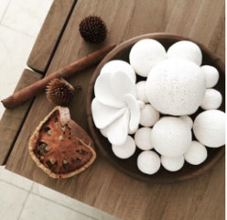 May include: A wooden bowl filled with white ceramic balls of various sizes. The bowl is on a wooden table with a cinnamon stick, dried fruit, and small brown pinecones.