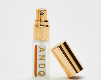 Puede incluir: Una botella de vidrio transparente y dorada con una tapa dorada y una etiqueta que dice "ANOQ". La botella está sobre una superficie blanca.