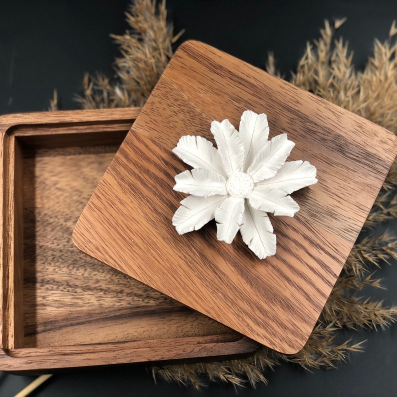 May include: A wooden box with a lid featuring a white flower design. The box is made of dark wood with a natural grain pattern.