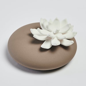 Può includere: Un diffusore in ceramica marrone a forma di pietra piatta con un fiore in ceramica bianca in cima.