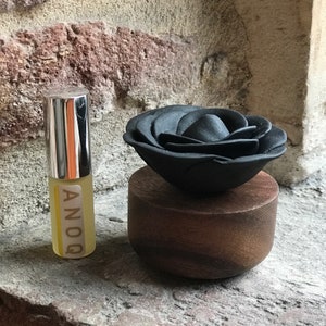 May include: A black rose diffuser sits on a wooden base. The diffuser is made of black ceramic and has a delicate, layered design. The base is made of dark wood and has a smooth, polished finish. The diffuser is labelled "ANOOQ".