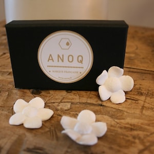 Op de afbeelding: Een zwarte doos met een goud en wit cirkelvormig logo en drie witte bloemvormige decoraties. Het logo luidt "ANOQ - MARQUE FRANCAISE". De items staan op een houten oppervlak.