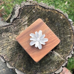 May include: A white ceramic flower with delicate petals sits on a square wooden coaster. The coaster has a natural wood grain and is a light brown color.